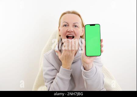 a woman opened her mouth and touched her chin with her hand she holds green screen chromakey phone in a tracksuit on an armchair with a plaid real people joy surprise good delivery discount housewife Stock Photo