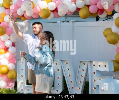 Just engaged couple raising a glass of champaign to their family standing under balloons and in front of a love sign after she said yes. Stock Photo