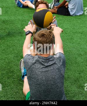 Two boys throwing a medicine ball to each other while doing situps on a turf field at sports summer camp. Stock Photo