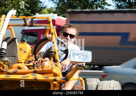 Smiling male tourist on vacation in the summer, with sunglasses, holding the wheel with a small homemade buggies, sitting in it parked on the side Stock Photo