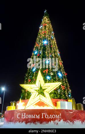 A large Christmas tree with bright garlands and star Shine lights down on the street in winter in the snow with the words 'happy New Year'. Decoration. Stock Photo