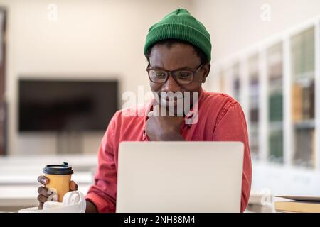 Satisfied hipster guy sit with coffee cup at table look at laptop screen read good news or message  Stock Photo