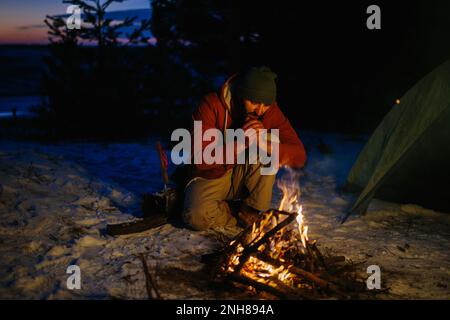 A man sits near a campfire, warming himself by the fire in a winter forest at sunset. Stock Photo