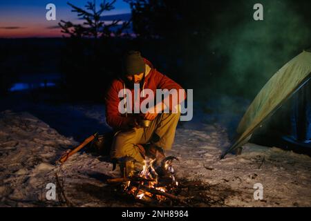 Image of hiker young man cooking dinner by a campfire in the mountains in winter. Male hiker with a beard sitting by the tent after sunset, making a c Stock Photo