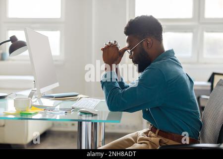 Thoughtful black male entrepreneur in glasses sitting at desk in front of computer, working online in office, side view Stock Photo