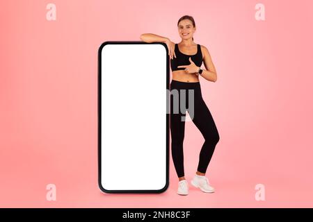 Mobile Mockup. Smiling Sporty Lady Pointing At Big Blank Smartphone Screen Stock Photo