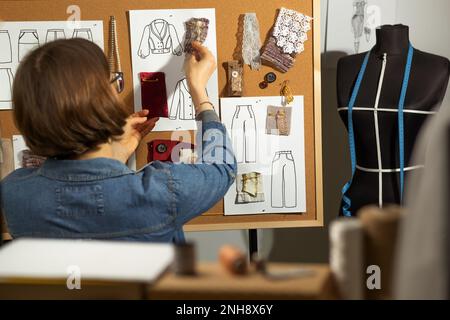 A woman designer works with sketches and samples of fabric for sewing clothes on a cork board. Stock Photo