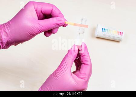 gloved hands holding glass tube with liquid and pink litmus test with acid pH level over light desk Stock Photo