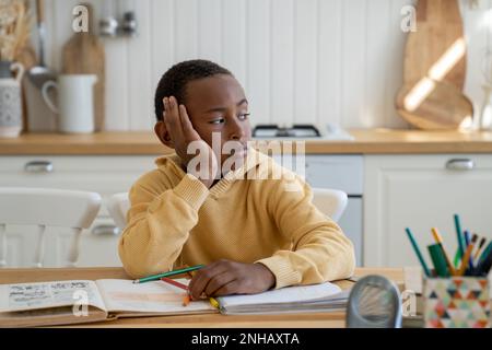 Thoughtful bored Black schoolboy look to side doing homework sits at table with textbooks at home Stock Photo