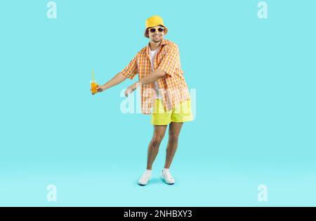 Happy young Caucasian man tourist performs dance with glass of juice in hands, stands in studio Stock Photo