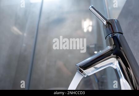 Water flowing from the chrome faucet on the sink in the bathroom. Shot showing the subject from the side, natural lighting conditions. Stock Photo