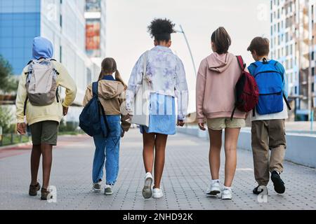 Rear view of youthful interracial pupils of secondary school with backpacks walking down street while coming back home after lessons Stock Photo