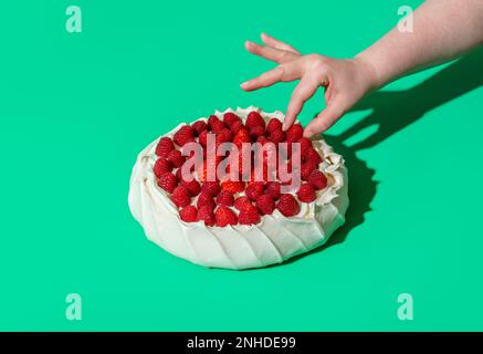 Woman's hand grabbing a raspberry from a pavlova cake, minimalist on a green background. Homemade pavlova cake with whipped cream filling, strawberrie Stock Photo