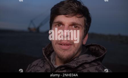Young Man in Work Gear Conducting Video Chat Against Backdrop of Construction Machinery During Evening Hours for Employer or Supervisor, Industrial Jo Stock Photo