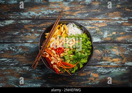 Asian, Chinese or Vietnamese style noodles salad with fresh vegetables, fried tofu and peanuts, served in rustic ceramic bowl on colorful wooden Stock Photo