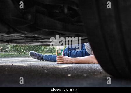 A mechanic on the ground, we can see his leg and arm holding a product. The picture has been taken underneath the car which permits to see one wheel. Stock Photo