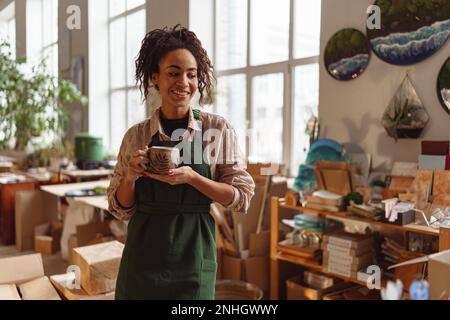 Woman florist decorator wearing apron drinking coffee during break in art studio Stock Photo