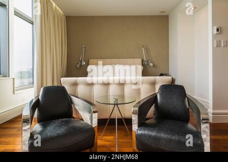 Black leather and chrome metal Club armchairs plus queen size sleigh bed with upholstered white cloth headboard and footboard in master bedroom. Stock Photo