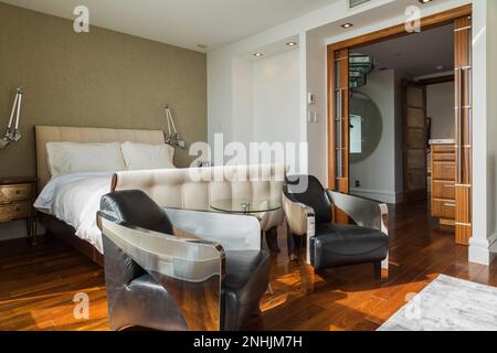 Black leather and chrome metal Club armchairs plus queen size sleigh bed with upholstered white cloth headboard and footboard in master bedroom. Stock Photo