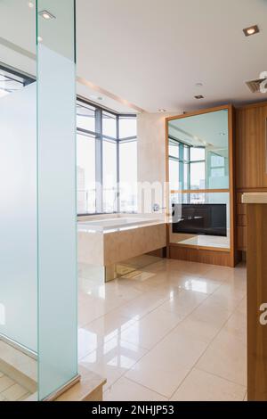 Glass shower stall, white bathtub with marble casing plus mirror with built-in wide screen television in en suite with marble tile floor inside condo. Stock Photo