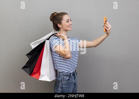 Side view portrait of woman wearing striped T-shirt broadcasting livestream and boasting her purchase, holding shopping, using smartphone. Indoor studio shot isolated on gray background. Stock Photo