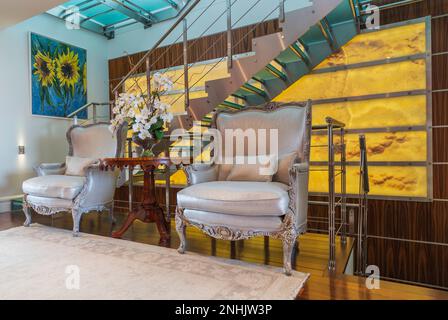 Wood and silver grey silk upholstered armchairs and steel and tempered glass staircase against exotic wood framed backlit nuanced backdrop in condo. Stock Photo