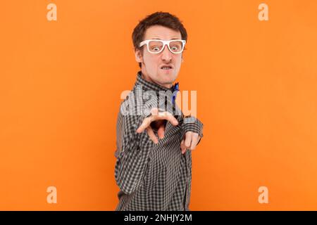 Portrait of funny crazy disabled man nerd pointing and looking at camera with clenched teeth, wearing shirt with blue bow tie and white glasses. Indoor studio shot isolated on orange background. Stock Photo