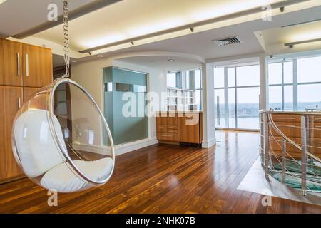 Clear plastic half-sphere chair suspended from the ceiling in home office with exotic wood cabinets and floorboards on top floor inside condo unit. Stock Photo