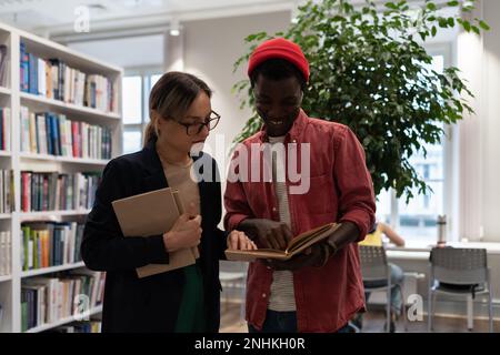 Focused professor woman helping understand textbook task to student African American man at library. Stock Photo
