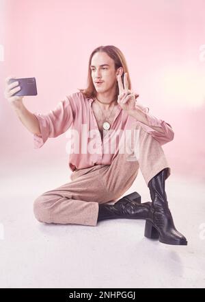 Selfie, peace and fashion with a gay man in studio on a pink background for lgbt inclusion or pride. Transgender, non binary and photograph with a Stock Photo