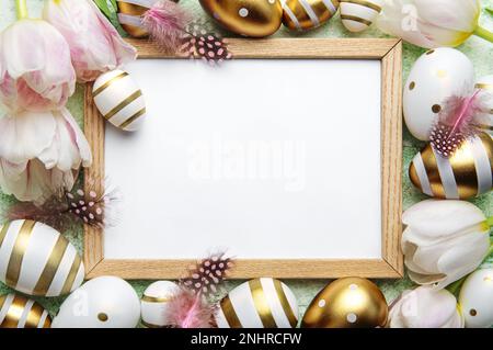 Blank picture frame surrounded by Easter eggs and tulip flowers on green background. Top view, flat lay composition Stock Photo