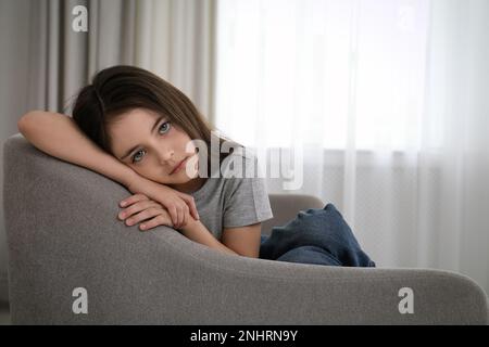 Sad little girl sitting on sofa indoors, space for text Stock Photo