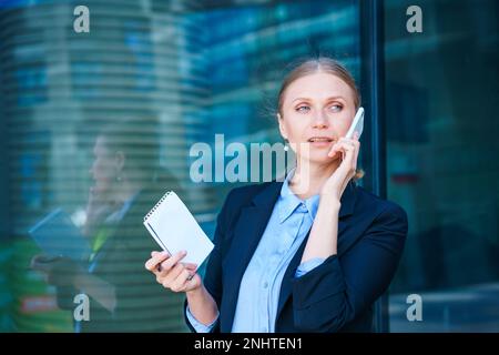 Woman in business suit carries folder with documents and smiles while talking on the phone. Business concept. Against the backdrop of an office building on a city street during the day Stock Photo