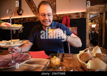 Smiling man drinking juice while sitting at a decorated breakfast table with a selection of cold cuts and healthy salads in foreground Stock Photo