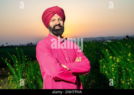 Portrait of happy young punjabi sikh man farmer standing cross arms wearing red turban and kurta looking at camera at agriculture field. Stock Photo