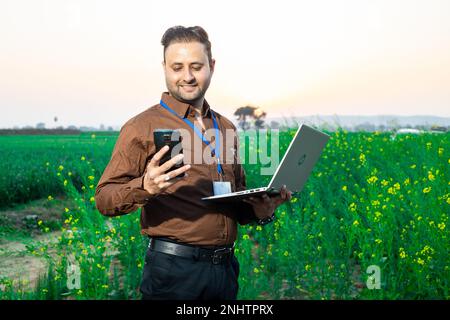 Portrait of happy young indian agronomist or government bank person holding laptop and using smart phone standing at agriculture field. Rural india. Stock Photo