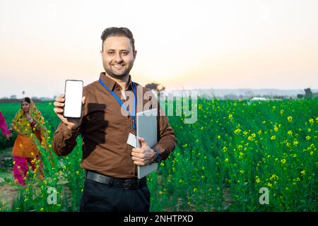 Portrait of happy young indian agronomist or government bank person showing smart phone with blank display to put advertisement standing at agricultur Stock Photo
