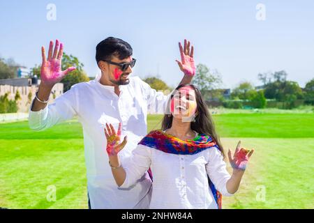 Cheerful young Indian couple wearing white kurta dancing celebrating celebrating holi festival at park, face painted with colorful powder or gulal. Stock Photo