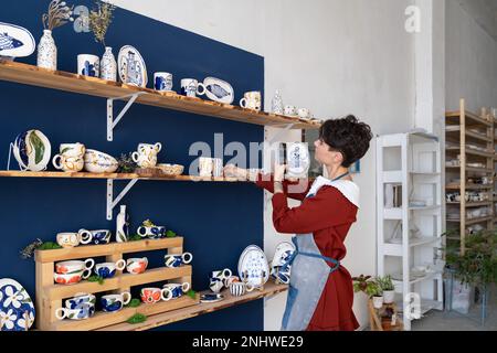 Entrepreneur craftswoman stands in own souvenir shop and takes pictures of handmade dishes on phone Stock Photo