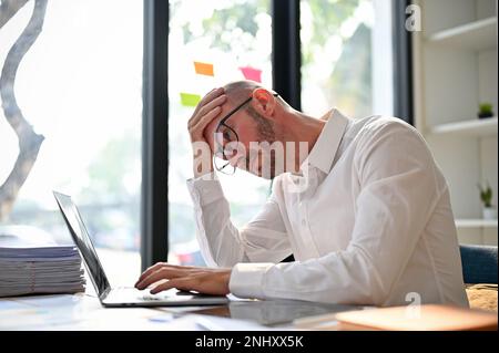 Stressed and unhappy caucasian businessman or male financial consultant having problems on his business, using laptop computer at his desk. overworked Stock Photo