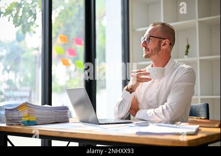 Smart and thoughtful caucasian businessman looking out the window, daydreaming about his successful career while sipping morning coffee at his desk. Stock Photo