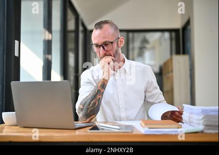 Professional and focused caucasian businessman with tattoo on his arm is working on financial data on laptop computer at his desk in the office. Stock Photo