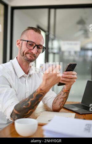 Thoughtful Caucasian man with glasses and tattoo on his arms holding a credit card or debit card and his smartphone at his desk. Stock Photo