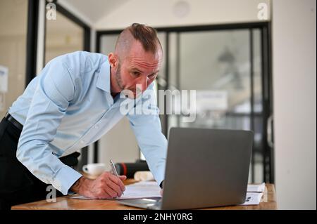 Smart Caucasian male engineer or contractor leaning on table, using laptop, noting something on paper, working on the building plan in the office. Stock Photo