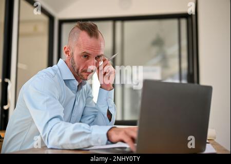 Thoughtful Caucasian male engineer or architect looking at laptop screen, pensive thinking and planning his work, focusing on a building plan on lapto Stock Photo