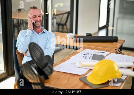 Happy and chilling Caucasian male engineer is on the phone with someone and putting his legs on the table while resting in the office. Stock Photo