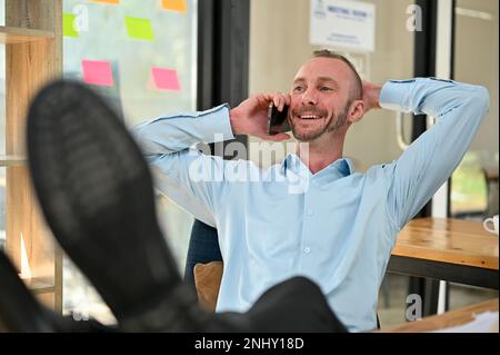 Handsome and happy Caucasian male engineer putting his legs on table and hand behind head while talking on the phone with client and chilling in the o Stock Photo