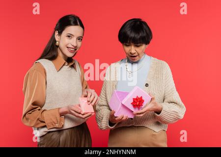 surprised asian woman opening gift box near young daughter smiling at camera isolated on coral Stock Photo