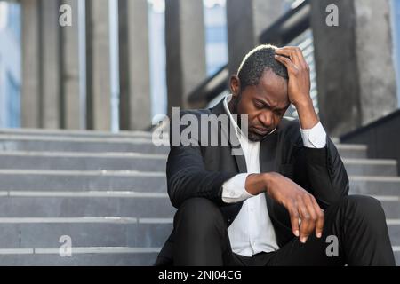 Sad african american businessman sitting on stairs outside office building, man in business suit bankrupt fired from job, boss lost money. Stock Photo