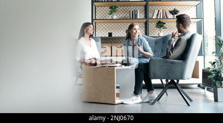 Interested happy colleagues sitting on chairs, listening to boss team leader, explaining working issues at meeting Stock Photo